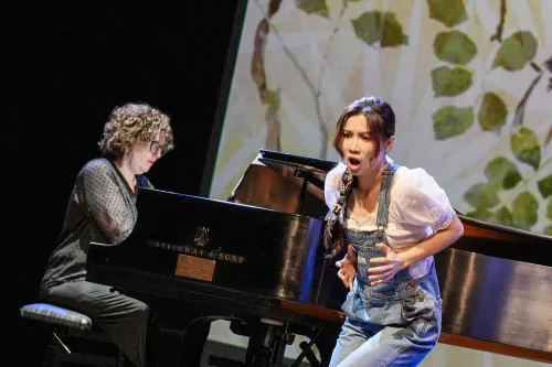 A woman in overalls leans forward and clutches her sides as she performs. 