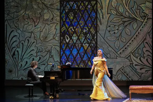 a woman in a gold ballgown peforms on stage with a pianist behind her.  The background is a projection of stained glass 