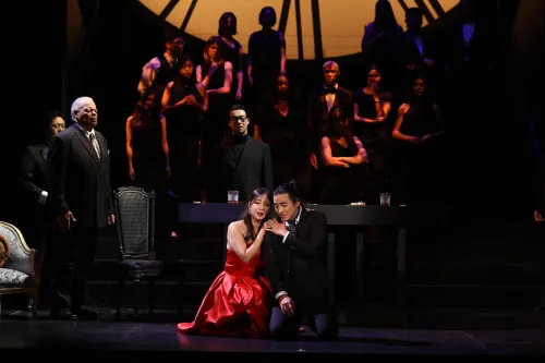 A man in a black suit and a woman in a red ballgown kneel and embrace on another, center stage, with a chorus of singers behind them.