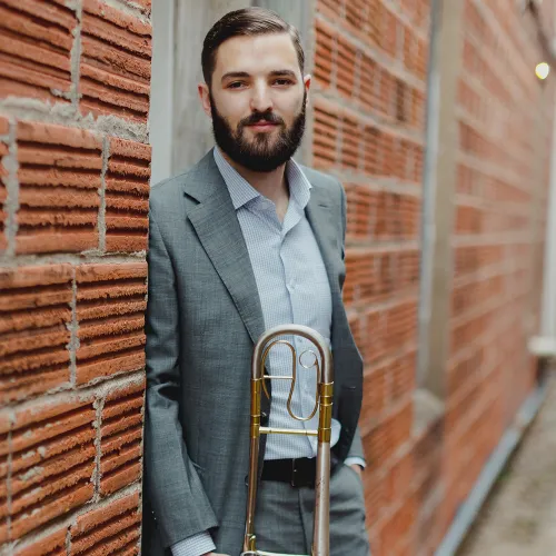 Sam Dimov stands against an industrial brick building, wearing a grey suit, holding his trombone