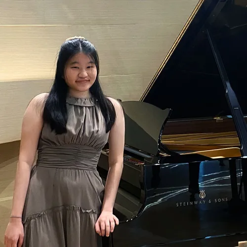 Kelly Chanpong stands at a grand piano and looks happily toward the viewer
