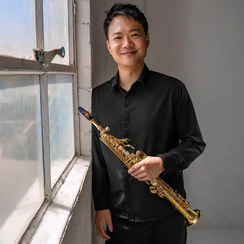 Calvin Wong stands at a window in a brightly lit room, holding a soprano saxophone and looking warmly toward the viewer. 