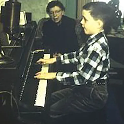 A very young Gregory Allen plays an upright piano while a woman looks on. 