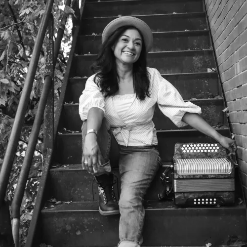 Susan Nevarez-Torres poses with her accordion on an outside staircase surrounded by trees. 