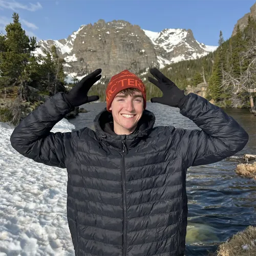 Connor McManus stands with his arms lifted to the sky, in front of an alpine lake with a snowcapped mountain in the background. 
