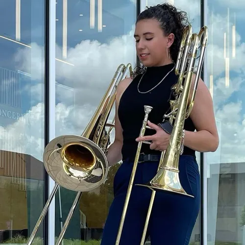 Jeanette Velasco stands infront of a bank of windows, looking down at a tenor and bass trombone she holds in each hand. 