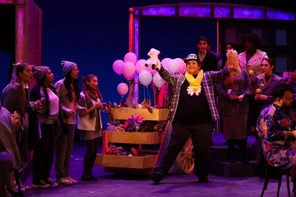 A man in a baseball cap and yellow scarf performs a gleeful song and dance number while other performers watch nearby. 