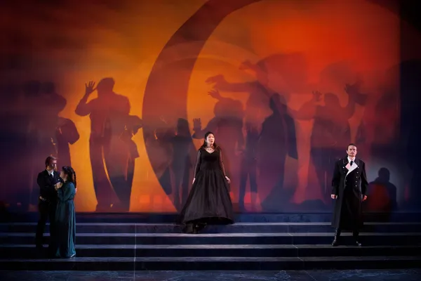 Three performers stand, spread across a wide, smoke-filled staircase, behind them is a spooky orange and red background with shadowy human figures projected against it. 
