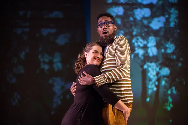 A man and woman embrace each other center stage in front of a forest projected onto the background of the stage.
