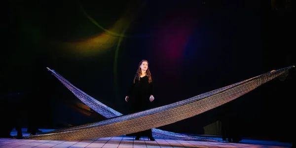 a performer in a black gown stand center stage, long strips of fabric cross-cross in front of her and behind her.