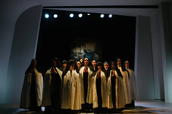 a group of nuns huddle together in dark light, hands clasped in prayer, looking up to the heavens.