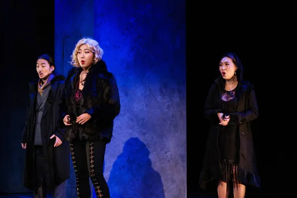 Three performers sing in a triangle formation, against a stark set, washed in lush blue light.