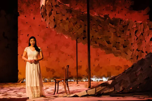 A woman in a white dress stands on a stage covered in paper, lit up in muted red tones.