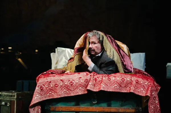 a man in a suit and tie lies on a bed, covered loosely by a comforter, his head is at the foot of the bed, and he peers out from the comforter as though he is hiding from someone. 