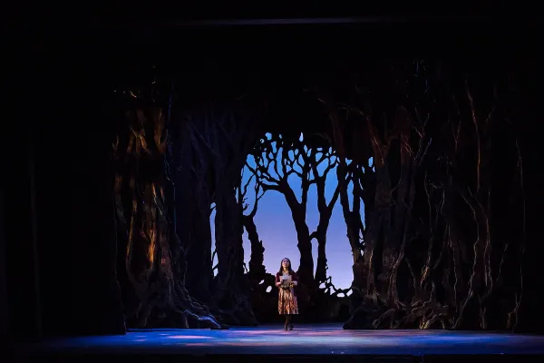 Gretel stands amid an epic forest set, holding a map and looking uncertain and frightened.