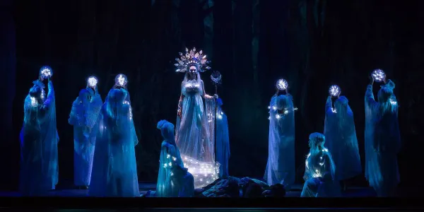 a witch stands on stage in ornate dress surrounded by shadowy figures.