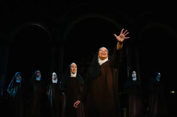 A nun stands center stage in a pool of light, with her arm stretched out in front of her, lifted hopefully to the sky, as several other nuns stand reverently behind her, fading in the darkness.