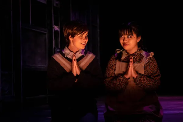 two children kneeled as if to pray, but they look knowingly at each other instead of praying.