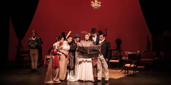 A group of people in formal Elizabethan attire stand center stage, huddled over newspaper that one woman holds up for all.