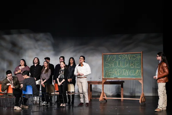 a group of singers stand in a row in a classroom as if at attention. Behind them a chalkboard reads "No Spanish should be heard in class."