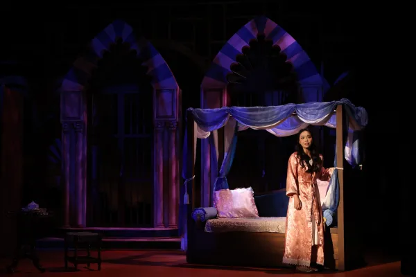A woman in a nightgown stands holding a bedpost in a dimly lit bedroom set, she is singularly lit with a spotlight. 