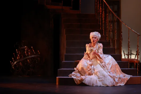 Marie Antoinette sits along on stage in an ornate ballgown and wig, looking somber.