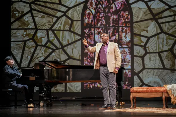 A man in a casual sport coat performs on stage, with stained glass projections behind him. 