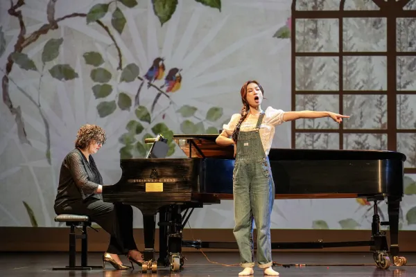 A woman in overalls stands in front of a grand piano on stage, pointing off to the left of stage as she sings.