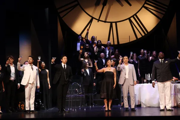 The cast and chorus of La Traviata stand together on stage, dressed in formal 1960s attire, with martini glasses outstreched toward the audience, in a group toast 