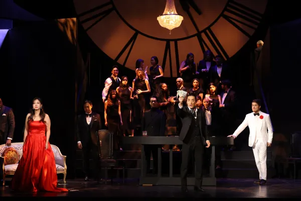 A man in a white suit and woman in a red ballgown stand on opposite ends of a stage, with a chorus of singers behind them. 