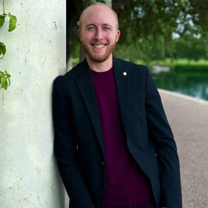 a headshot of Micah Rosenstien, leaning against a white pillar, surrounded by lush greenery