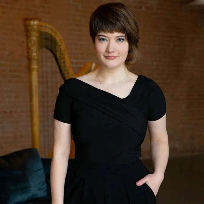 Emily Levin stands in front of her harp and looks toward the viewer
