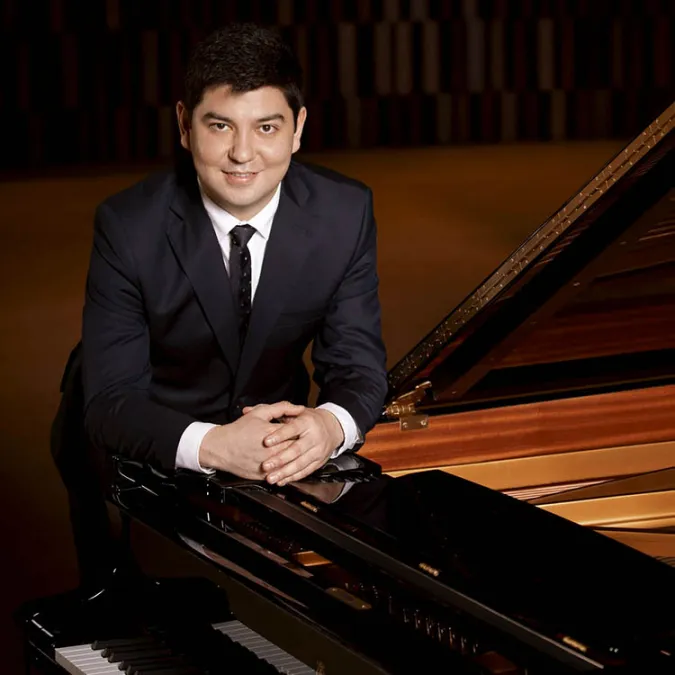 a headshot of Behzod Abduraimov in formal attire leaning against a concert grand piano with his arms folded.