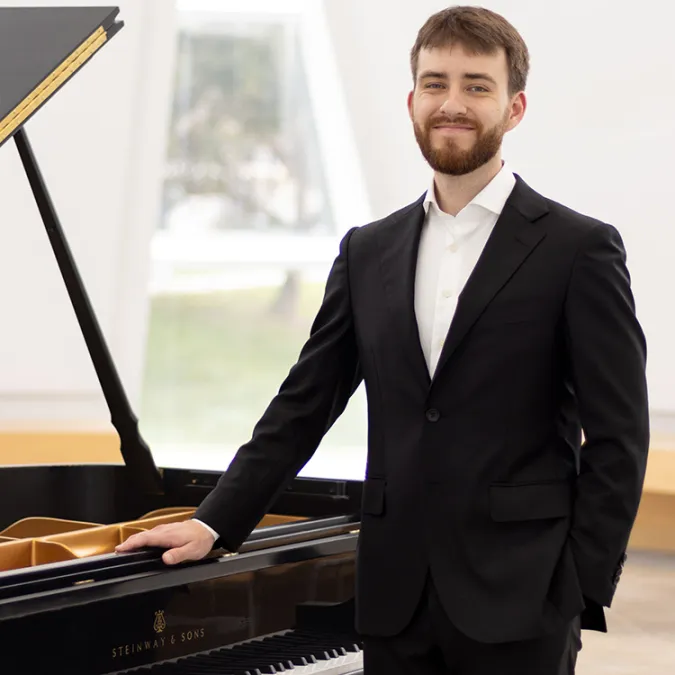Ryan Aguilar stands near a grand piano in a brightly lit room.