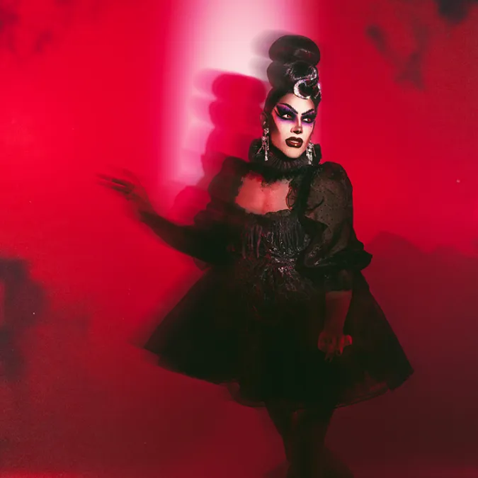 a publicity still of Evah Destruction, in gothic makeup and a black dress, standing against a red and black wall.