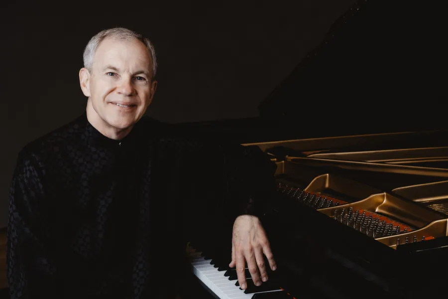 Anton Nel sits with his left arm resting on top of a grand piano wearing a black shirt.