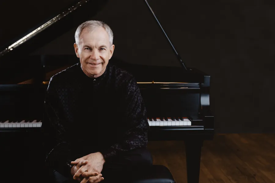 A man with gray hair wearing a black shirt smiles and sits in front of an open piano.