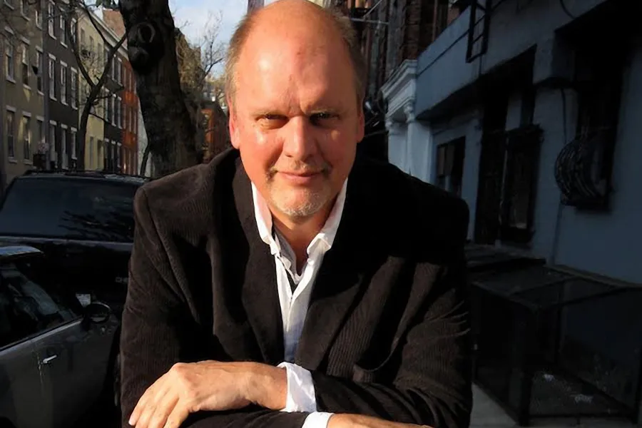 A white man in a white shirt and black jacket sits outside with his arms crossed and smiles.