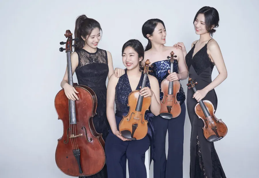 Four asian women hold their string quartet instruments and smile at one another. They are wearing formal concert attire.