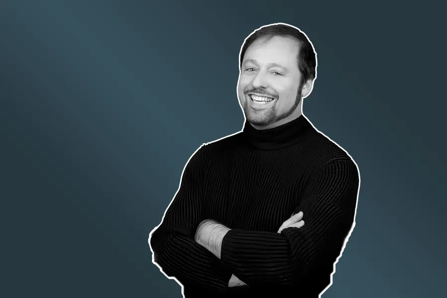 A black and white photo of pianist Steven Mayer with his arms crossed against a blue background.