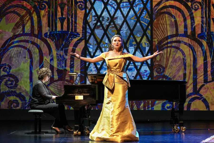 A singer in a formal yellow ball gown performs on stage with her arms outstretched, as a pianist accompanies behind her. 