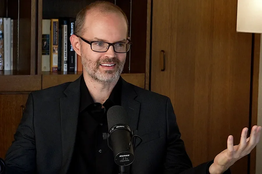 Chris Prosser Talks into a mic with a bookshelf in the background