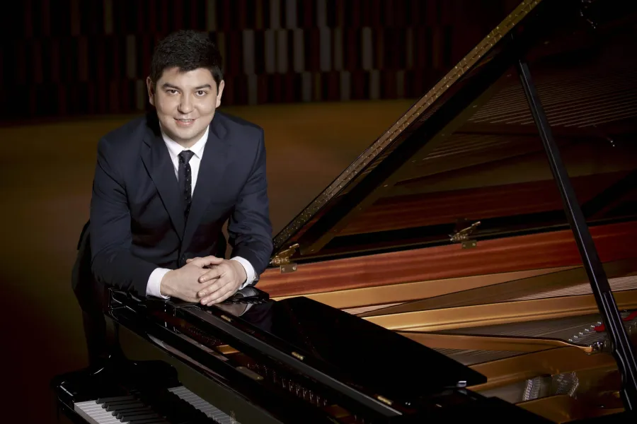 Pianist Behzod Abduraimov in a suit leaning on a piano