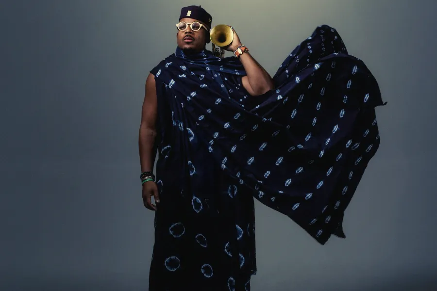Composer Etienne Charles, wearing a navy blue patterned kaftan and shawl, holds his trumpet beside his ear