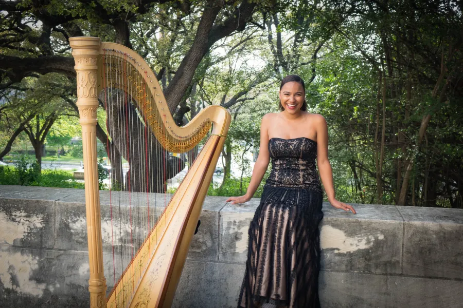Mallory McHenry leans on a stone wall wearing a black and gold strapless gown beside her harp.