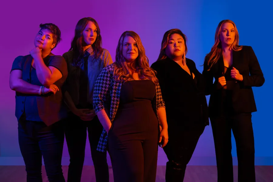 The members of VAMP dressed in black masculine clothing in front of a purple and blue background.