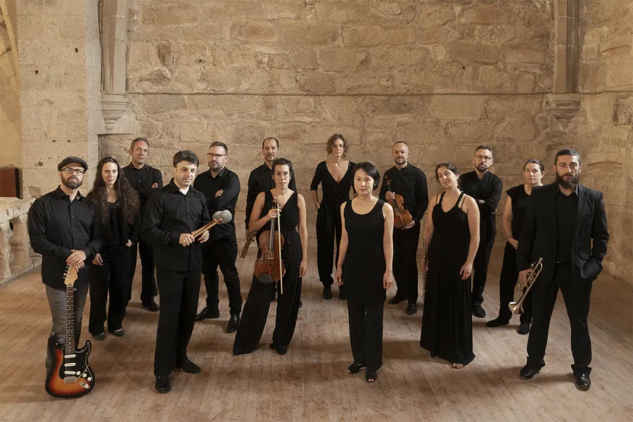 Members of Vertixe Sonora Ensemble stand in a stone room, dressed in black, holding their instruments.