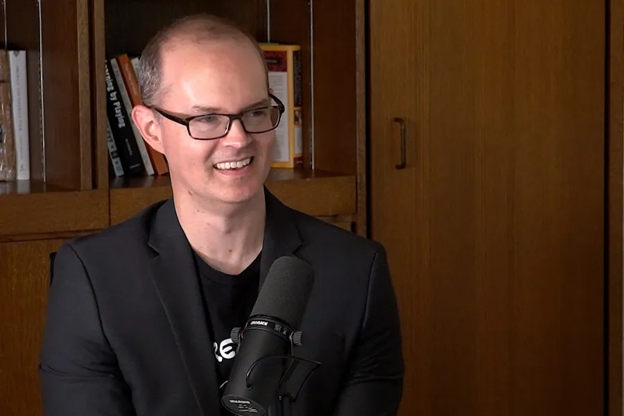 Chris Prosser Talks into a mic with a bookshelf in the background