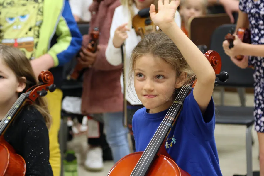 A child holds a cello in class and is raising their hand