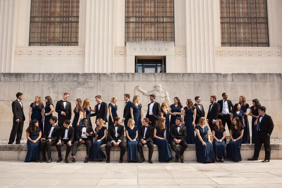 A large group of students dressed in formal attire posed casually in front of a University of Texas building. 